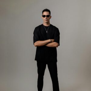 Stylish man in sunglasses and black attire standing confidently against a gray backdrop.